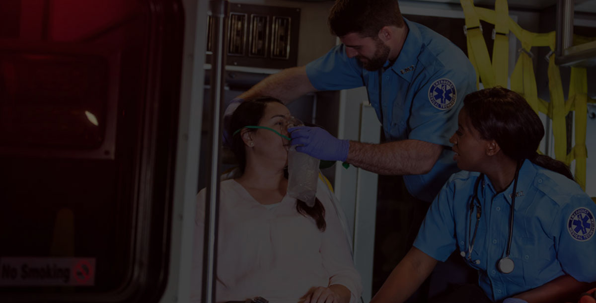 Two multi-ethnic paramedics, a Caucasian man and African-American woman, assisting a patient, giving her oxygen inside an ambulance. The patient, a mature Hispanic woman, looks worried.