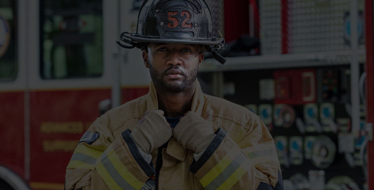 A firefighter standing in front of a fire truck, wearing a fire protection suit and helmet. He is a young African-American man in his 20s.