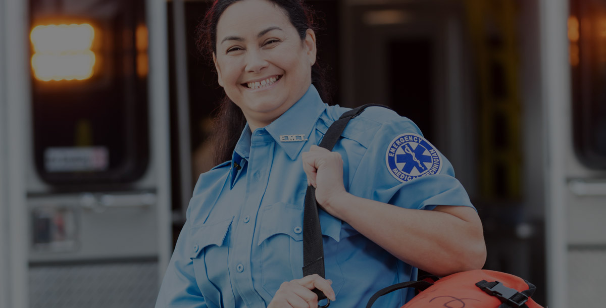 A mature Hispanic woman in her 40s, a paramedic, standing at the rear of an ambulance, by the open doors. She is looking at the camera with a confident expression, smiling, carrying a medical trauma bag on her shoulder.